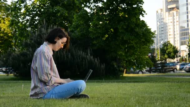 Young brunette woman sitting on green grass and use laptop — Vídeo de stock