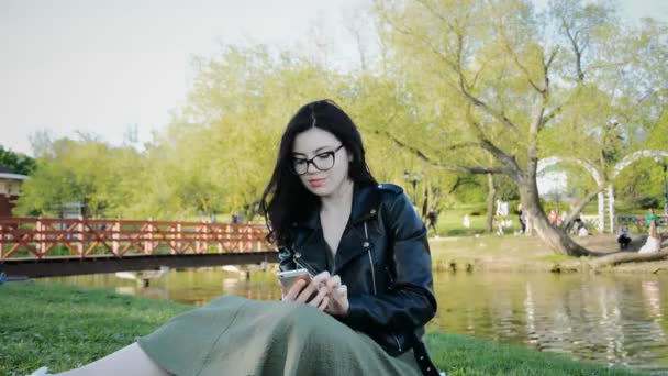 Młoda studentka korzystająca ze smartfona siedząc na zielonym trawniku — Wideo stockowe