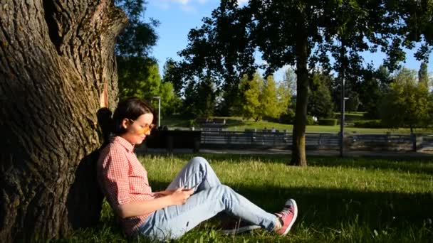 Una chica en un parque de verano se sienta en un césped cerca de un árbol y escribe un mensaje — Vídeo de stock