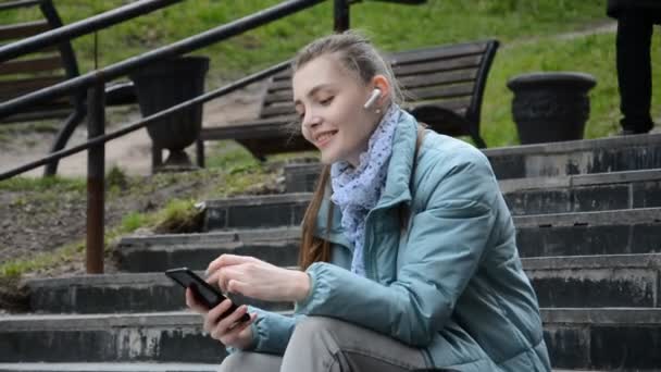 Seitenansicht der fröhlichen jungen Studentin, die auf den Stufen sitzt, hört Musik und lächelt mit — Stockvideo