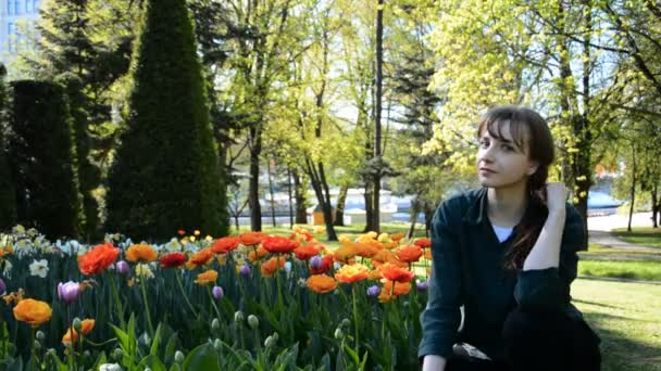 Una hermosa joven se sienta en un césped en un parque de verano junto a un club de tulipanes de colores — Vídeo de stock