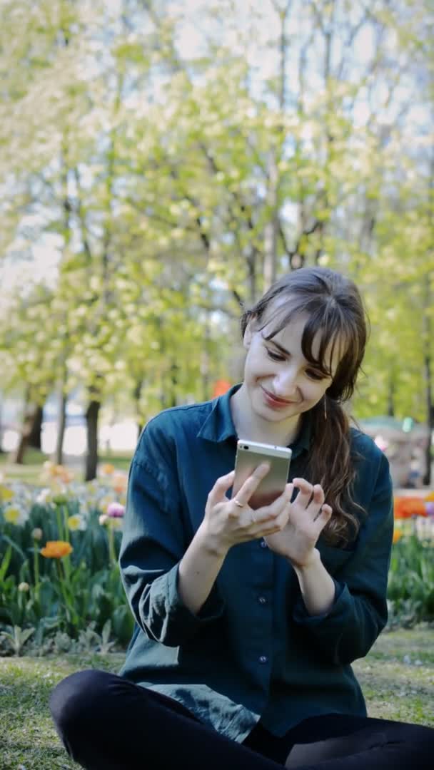 Una bella giovane ragazza è seduta su un prato in un parco cittadino estivo e chatta tramite uno smartphone — Video Stock