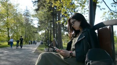 Bir yaz parkında bankta oturan esmer kadın internet üzerinden akıllı telefondan internet üzerinden ödemeler yapıyor.