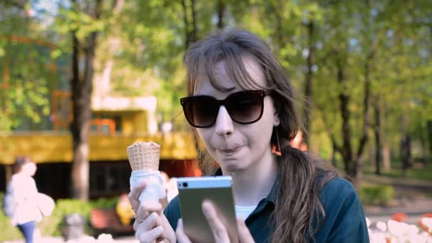 Junge schöne Frau im Sommer im Park isst leckeres Eis und liest die Nachrichten auf ihrem Handy — Stockvideo
