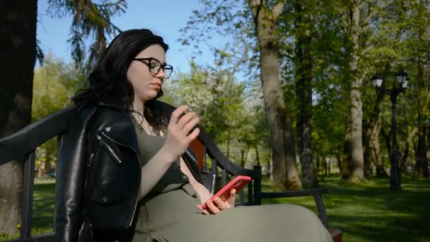 Joven hermosa chica está sentado en un banco en el parque y navegar por la cinta de redes sociales en su teléfono rojo — Vídeo de stock