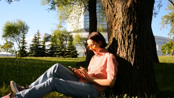 Młoda szczęśliwa kobieta korzysta z telefonu siedząc na trawniku w pobliżu nowoczesnego budynku miasta — Wideo stockowe