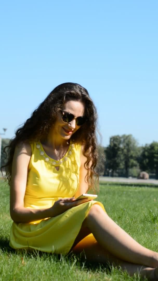 緑の草の上に座っている幸せな若い大人の女性は、電話でメッセージを入力しながら — ストック動画