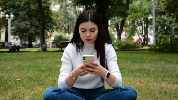 在夏天的公园里，一个年轻的女人坐在绿色的草坪上，手里拿着智能手机 — 图库视频影像