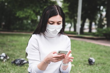 Koruyucu maskeli genç bir kız parkta dizüstü bilgisayarında ve elinde telefonuyla çalışıyor.