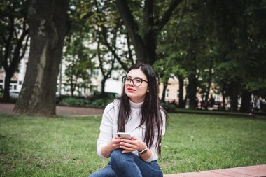 Kot pantolonlu güzel bir kız ve elinde telefonla parkta beyaz bir kazak.