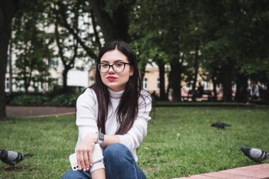 Kot pantolonlu güzel bir kız ve elinde telefonla parkta beyaz bir kazak.