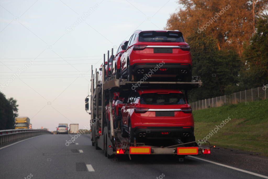 Two-level car carrier truck transports red chinese new cars on suburban highway road, back view, delivery autos logistics, export import automobile carriage transportation business