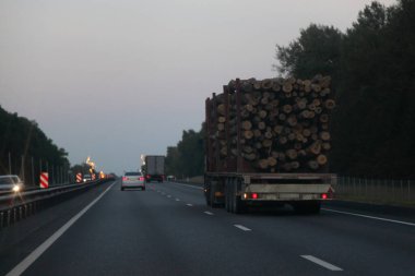 Yaz akşamı asfaltlanmış banliyö otoyoluna 3 eksenli karavan ile kereste kamyonu yüklenmiş. Arka plan manzarası, ormancılık endüstrisi kereste taşımacılığı.
