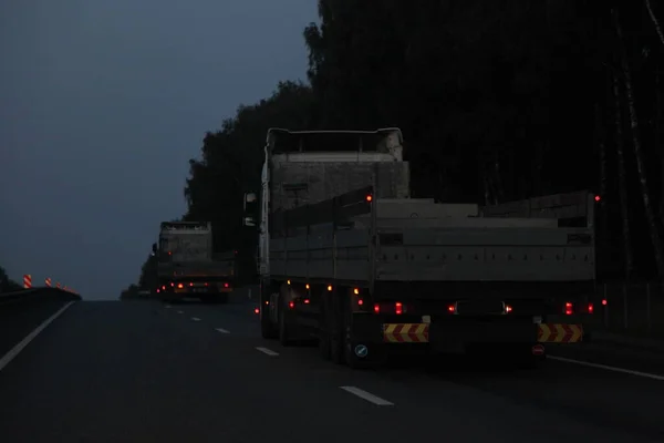 Loaded lowbed semi trailer trucks move on dry asphalted night road in dark, back view - intrenational transportation logistics