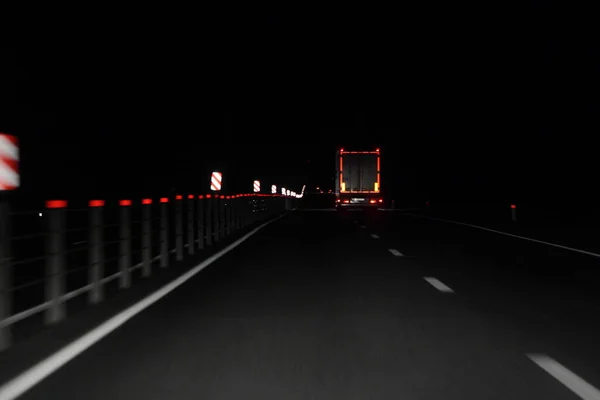 Suburban dry asphalted night road in dark with car head lamp light and back view moving Semi trailer truck with lighting reflection stripe on rear board  - night drive safety