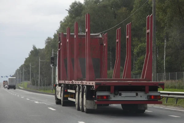 Empty wood truck transporter drive with a red trailer on a suburban asphalt highway road on summer day back side  view on green forest background, timber transport, forestry business