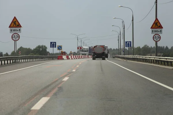 Narrowing road with road repair sign, road works on a suburban highway on a summer day, rear view on gray sky and milk truck with a gray barrel background