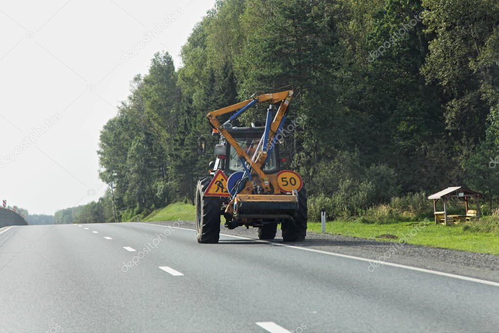 Wheeled tractor lawnmover with a external detachable mounted mower equipment movng on roadside of a suburban highway on forest background at summer day, road maintenance service mechanization machine