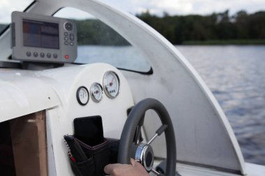 Yüzen motorlu tekne direksiyonunda bir adamın eli, kontrol aletleri ve harita çizicisiyle kabin iç gösterge paneli yaz günü su ve nehir kıyısı arka planında, açık hava seyahati