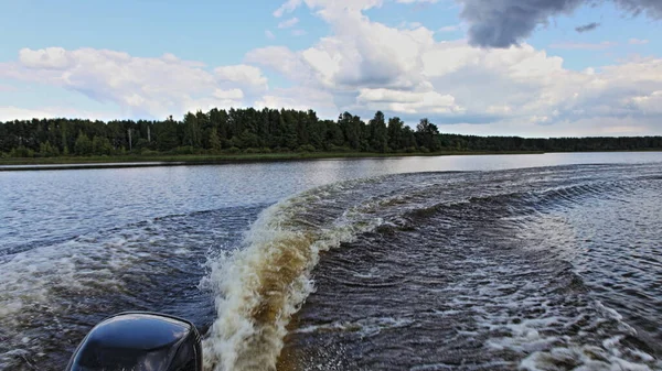 A speedboat turns on the river water with circle keelwater trail on green grassy bank background at summer day, rear view on foamy transom wake from stern of motor boat with single outboard motor
