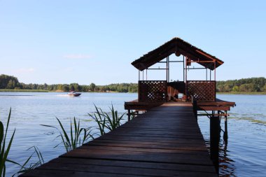Boş ahşap balkon cambaz ayaklıkları üzerinde tahta Pier köprüsünün perspektifi nehir suyuna motorlu tekne, mavi gökyüzü ve yeşil orman yaz günü ufukta, açık hava tatil nehri manzarası