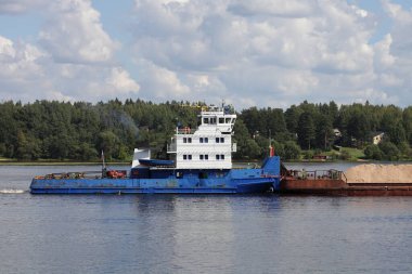 Rus satıcı mavi yük römorkörü, yeşil orman kıyısı ve mavi gökyüzü arka planında Volga nehrinde dolu bir mavna taşıyor. Nehir lojistiği, deniz yoluyla kum taşımacılığı. 