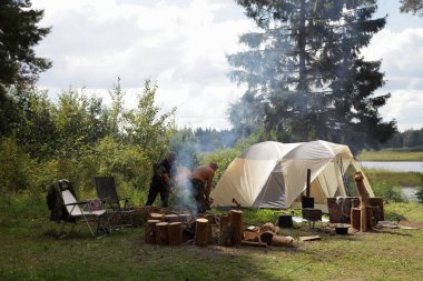 Turist kampı hayatı, kamp çadırında odun kesen iki izci, güneşli bir yaz gününde nehir kıyısındaki bir ormanda çam ağaçları, açık hava turizmi, romantik yaşam tarzı.