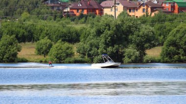 Bir kulesi olan eski bir iç çekme teknesi, Wakeboardcuyu çekiyordu. Yeşil nehir kıyısında yaz günü kır evi olan bir arka planda, nehirde aktif bir dinlenme, su kayağı, wakeboard.