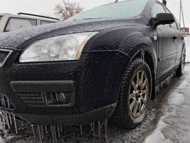 Ice-covered parked car front side view in perspective, headlight, wheel, bumper with icicles and windscreen, icy vehicle on a winter day after freezing rain in Europe clipart