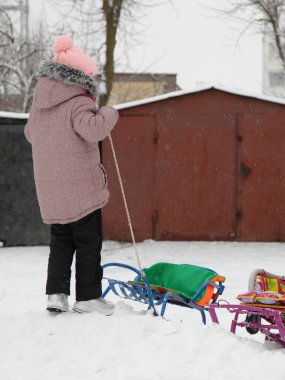 Bir kız çocuğu, bir kış günü tepede kar üzerinde bir halatı eski bir garajın arka planına doğru çeker.