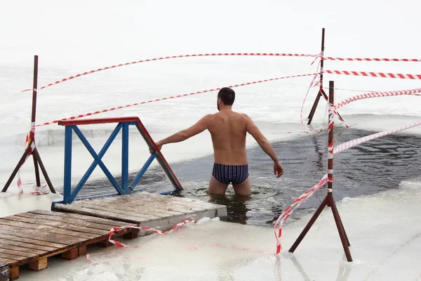 Winter swimming sport, a Caucasian man in a swimming trunks enters in the ice hole water on planked footway on a Sunny frosty winter day, healthy lifestyle