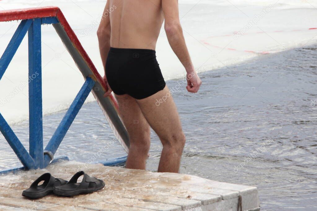 Winter swimming sport, a European guy in a swimming trunks enters in the ice hole water on planked footway on a Sunny frosty winter day, healthy lifestyle