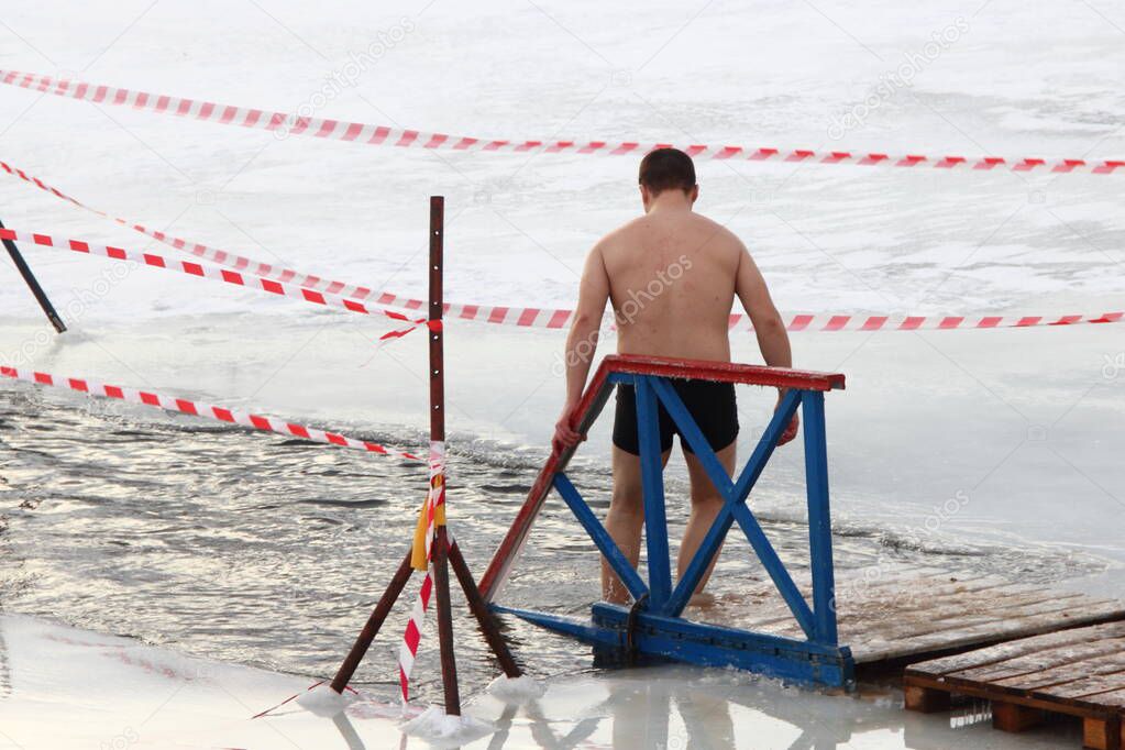 Winter swimming sport, a European man in a swimming trunks enters in the ice hole water on planked footway on a Sunny frosty winter day, healthy lifestyle