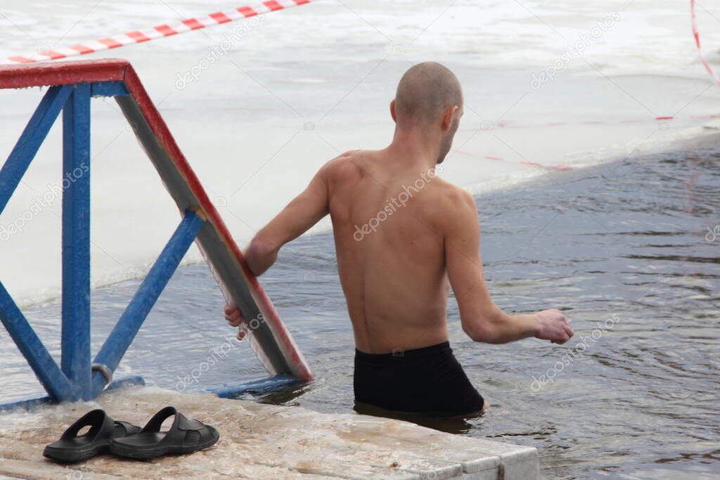 Winter swimming sport, a Caucasian hairless skinny boy in a swimming trunks enters in the ice hole water on planked footway on a Sunny frosty winter day, healthy lifestyle