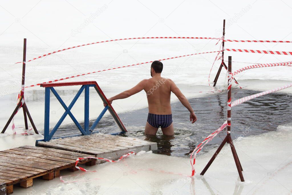 Winter swimming sport, a Caucasian man in a swimming trunks enters in the ice hole water on planked footway on a Sunny frosty winter day, healthy lifestyle