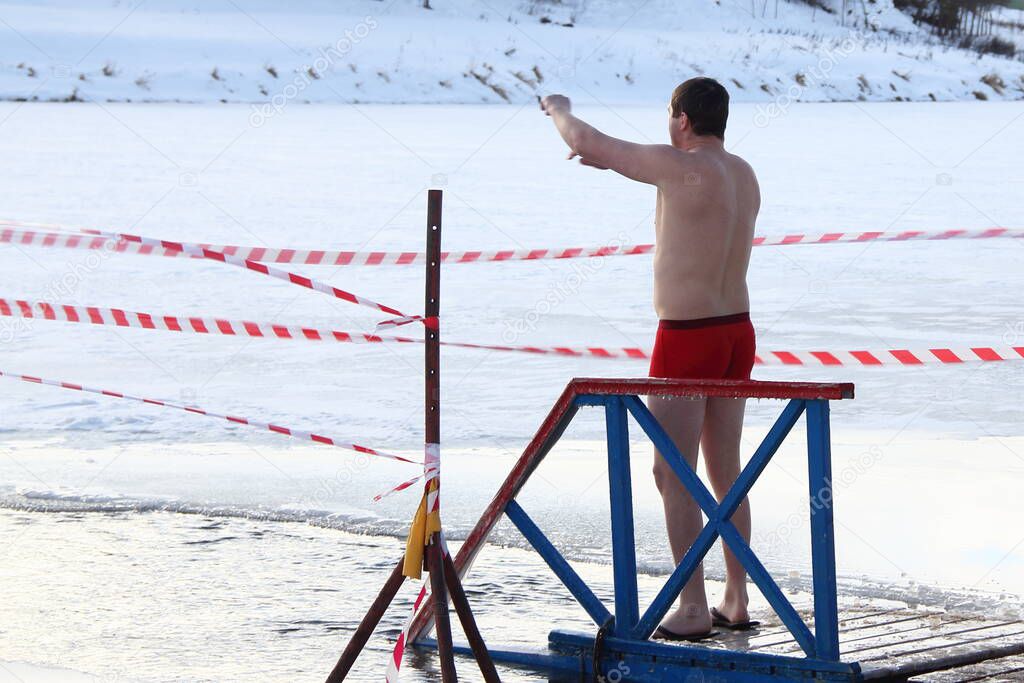 Winter swimming sport, a Caucasian man in a swimming trunks stand up near the ice hole water on planked footway on a Sunny frosty winter day, healthy lifestyle