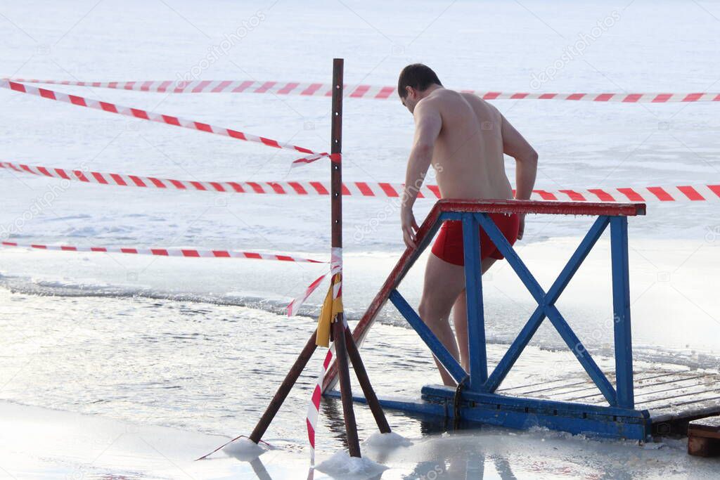 Winter ice swimming sport, a European man in a swimming trunks enters in the ice hole water on planked footway on a Sunny frosty winter day, healthy lifestyle