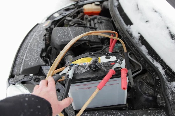 The hand connects the negative terminal on frozen car engine cold start with spare battery accumulator and wires with crocodile clips at winter day