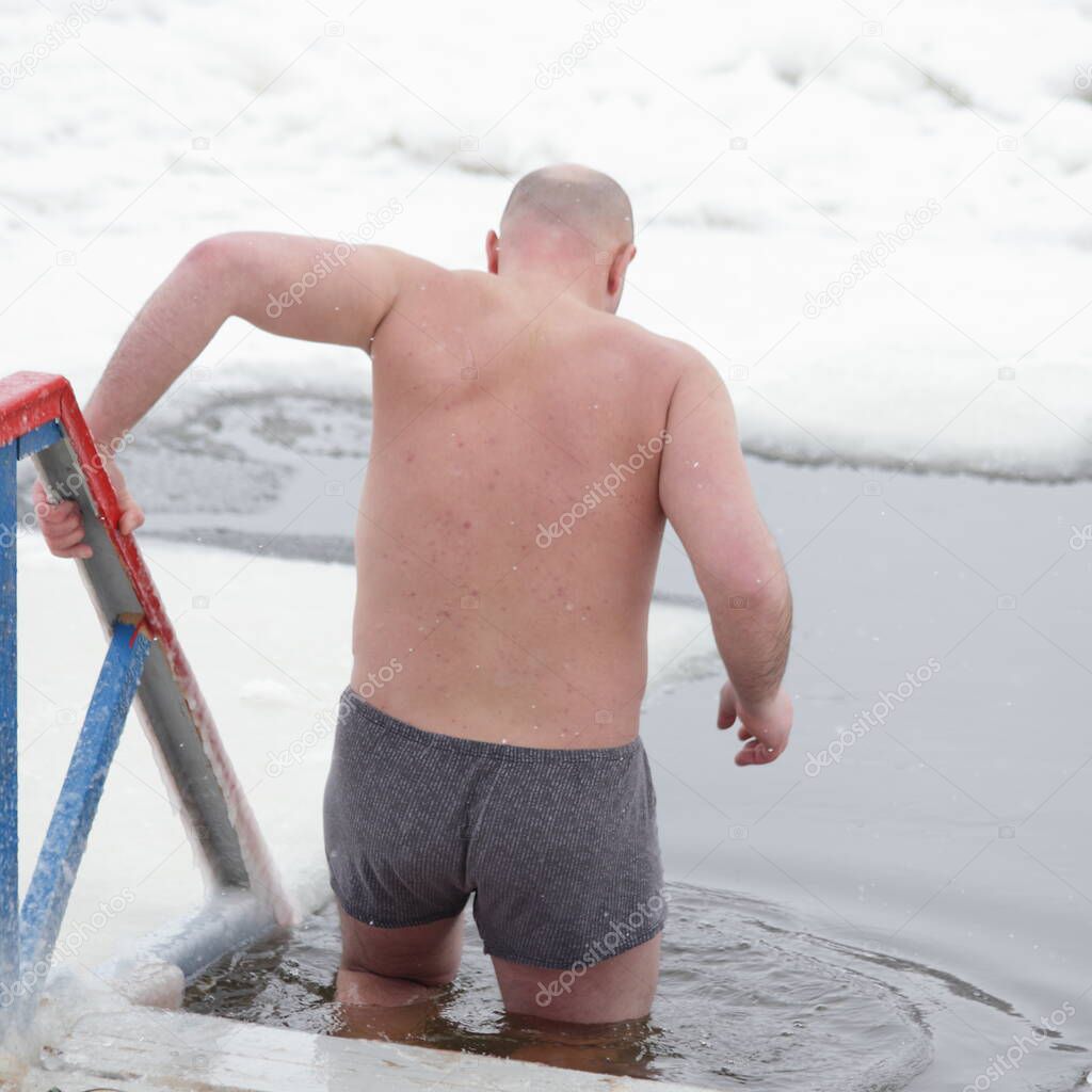 Winter ice swimming sport, a Caucasian man in a swimming trunks enters in the ice hole water on planked footway on a Sunny frosty winter day, healthy lifestyle