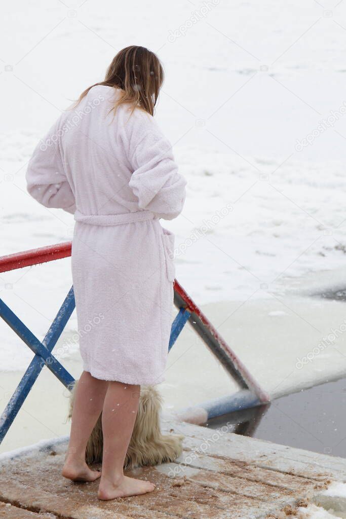 Winter swimming sport, a mature Caucasian woman in a in a white coat, getting ready for a swim the ice hole water on a Sunny frosty winter day, healthy lifestyle