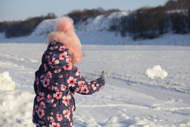 Kışlık giysiler içindeki bir çocuk, güzel banliyö kış buzlu nehrinin arka planına kartopu atar. Donmuş su ve kar kaplı kıyı tepesi güneşli kış günlerinde çıplak ağaçlarla kaplıdır.