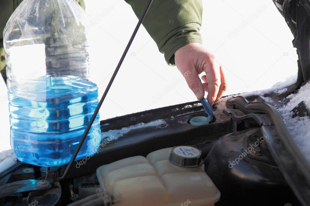 The driver's hands open the cap to fill up the non-freezing blue windshield washer fluid of the windshield washer in the tank of the car on a winter sunny day
