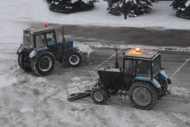 İki tekerlekli traktor turuncu lamba ile kar temizliyor kazıyıcı kürek ile yol kenarında süpermarket otoparkı yakınında kış akşamında yoğun kar yağışı sonrası, üst taraf manzarası