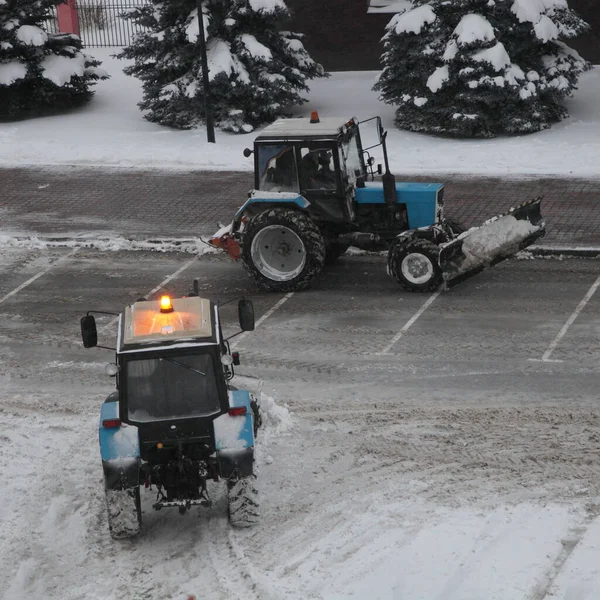 Two wheeled traktors  cleaning a snow with scraper shovel blade on road near supermarket parking place after heavy snowfall at winter evening, top side view