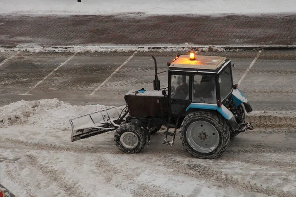 One wheeled traktor with orange signal lamp cleaning a snow with scraper shovel blade on road near supermarket parking place after heavy snowfall at winter evening, top side view