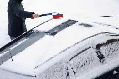 Kar kaplı arabanın çatısından kışın büyük saplı kırmızı fırçayla kar temizlerken, kar yağdıktan sonra araba sürmeye hazırlanıyor.