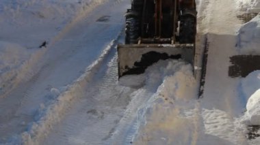 Büyük tekerlekli bir buldozer traktörü, kar küreğiyle kar temizliyor. Şehir otoparkında, kışın yoğun kar yağışı sonrası, üst tarafı kapatıyor.