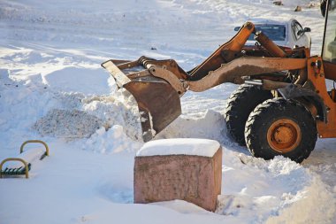 Sarı tekerlekli bir traktör, Sunny kış gününde yoğun kar yağışı sonrası kar kürekli kar küreme küreği ile kar küreme aracını parkın yanındaki bankın yanına kaldırır.