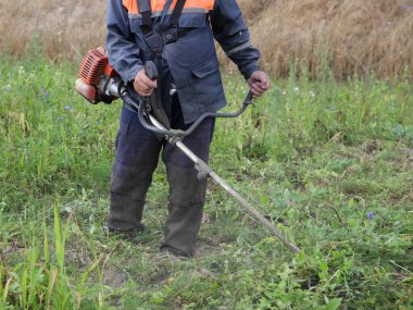 Belinde iş kıyafetleri olan, çim biçme makinesiyle çimleri biçen, yaz günü yeşil çimlerde çim biçen, bahçe aletlerinin önünü kapatan çiftçi.