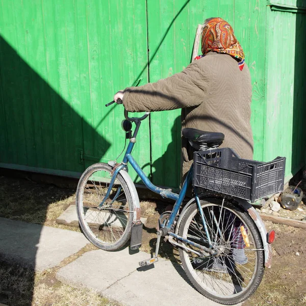 An elderly Russian woman in warm clothes carries a bicycle against the background of a wooden house in the village on a Sunny spring day, pensioner people rural transport